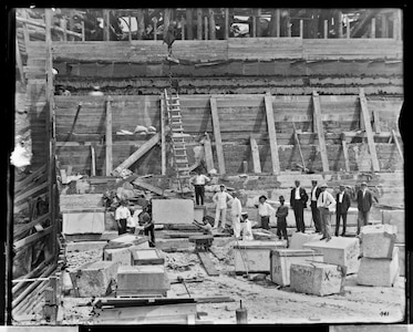 This photograph from August 23, 1905 shows the first granite block being lowered into Dry Dock 3 as naval officers, shipyard leadership, and personnel look on.