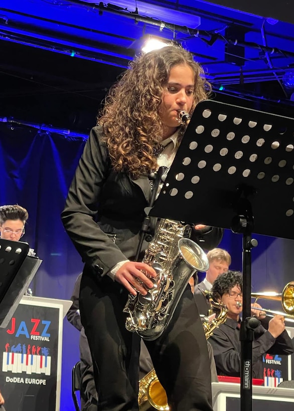 A young woman plays a sax on stage other members of her band are seated behind her.