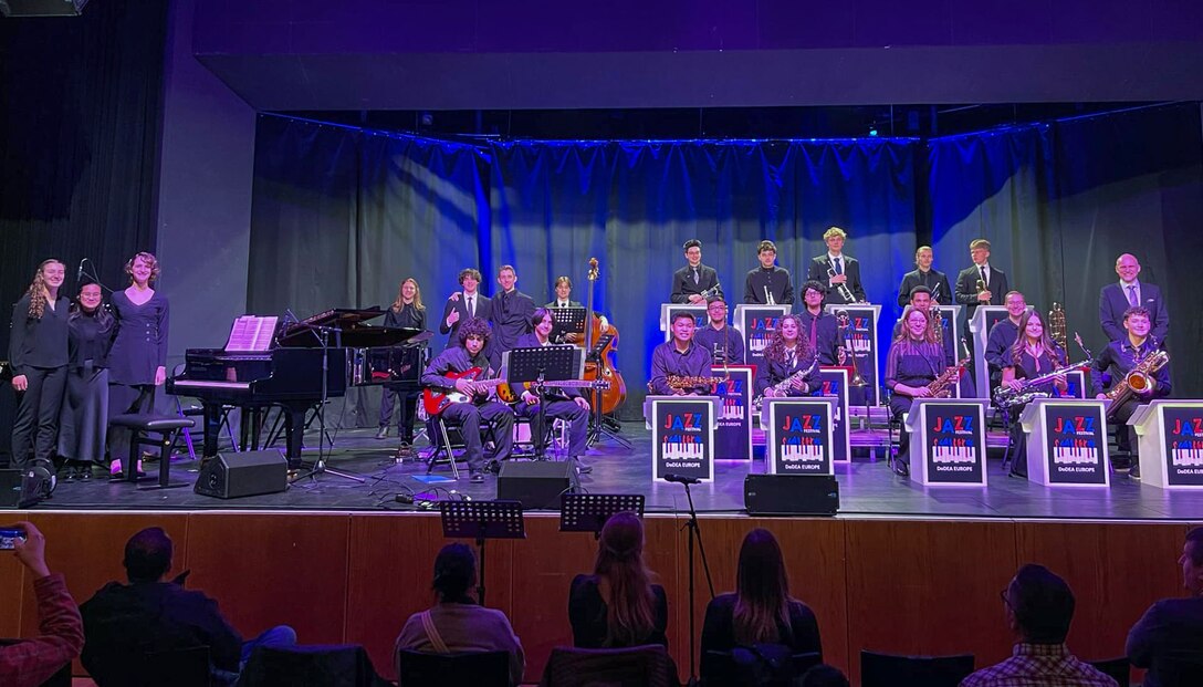 About 20 students and one adult stand on a stage after performing music. the audience in the foreground is applauding.
