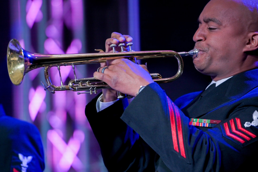 Close up photo of a man in a dark Navy uniform playing a trumpet on stage
