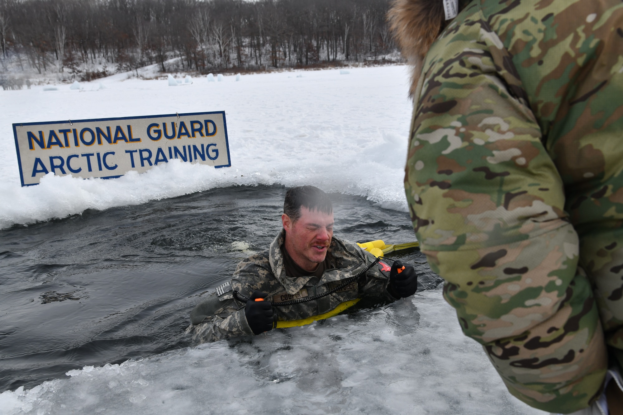 A man emerges from an icy pool of water carved into a snowy lake. A sign behind him reads “National Guard Arctic Training.”