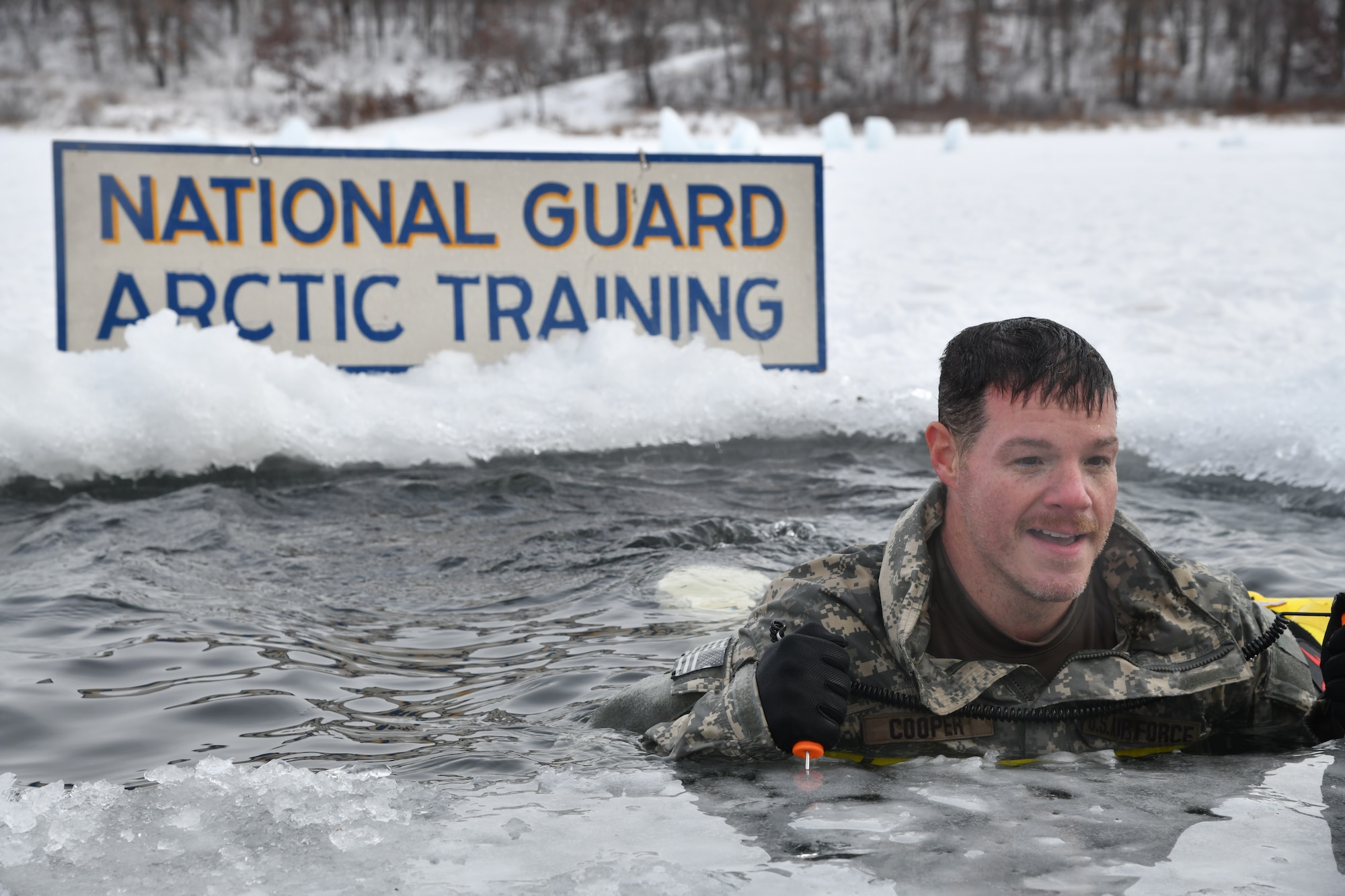 A man emerges from a icy pool carved into a snowy lake. A sign behind him read "National Guard Arctic Training."