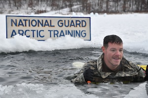 A man emerges from a icy pool carved into a snowy lake. A sign behind him read "National Guard Arctic Training."