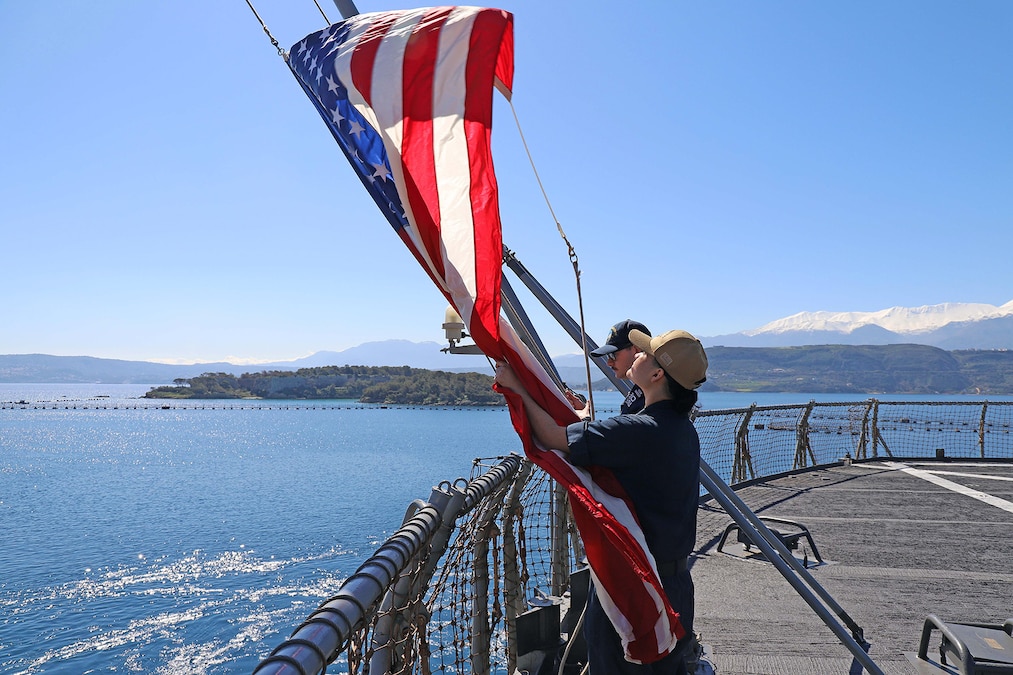 Two sailors raise the flag on the deck of a ship with islands in the distance.