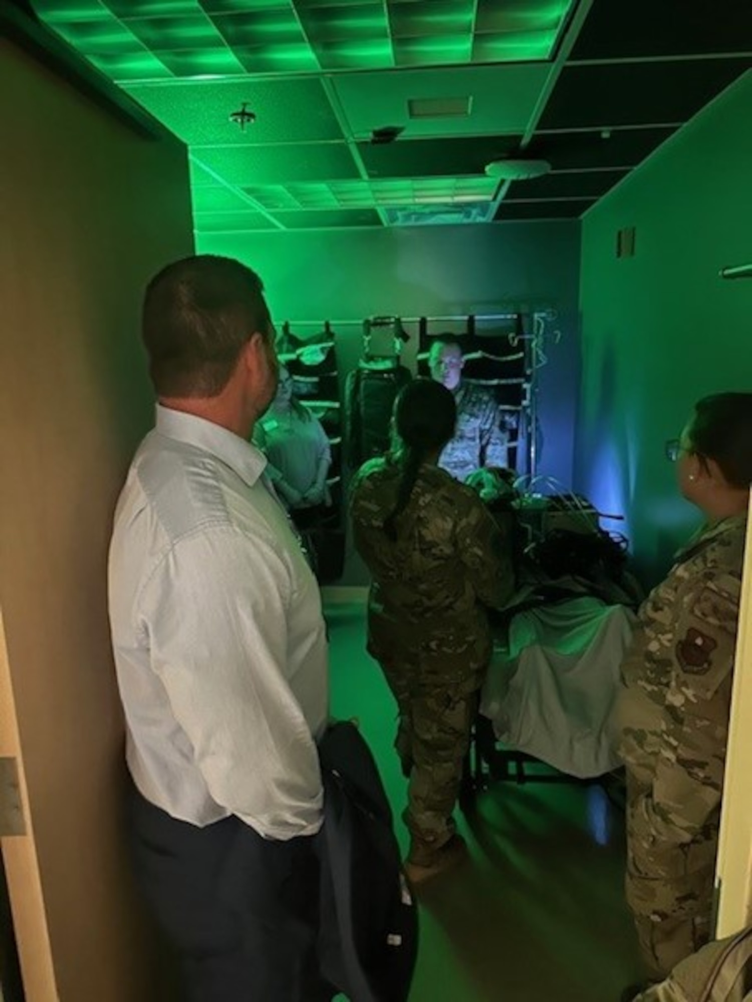 A man in business casual attire and three servicewomen look forward, towards a serviceman talking with a blue light shining near his face in a green room.