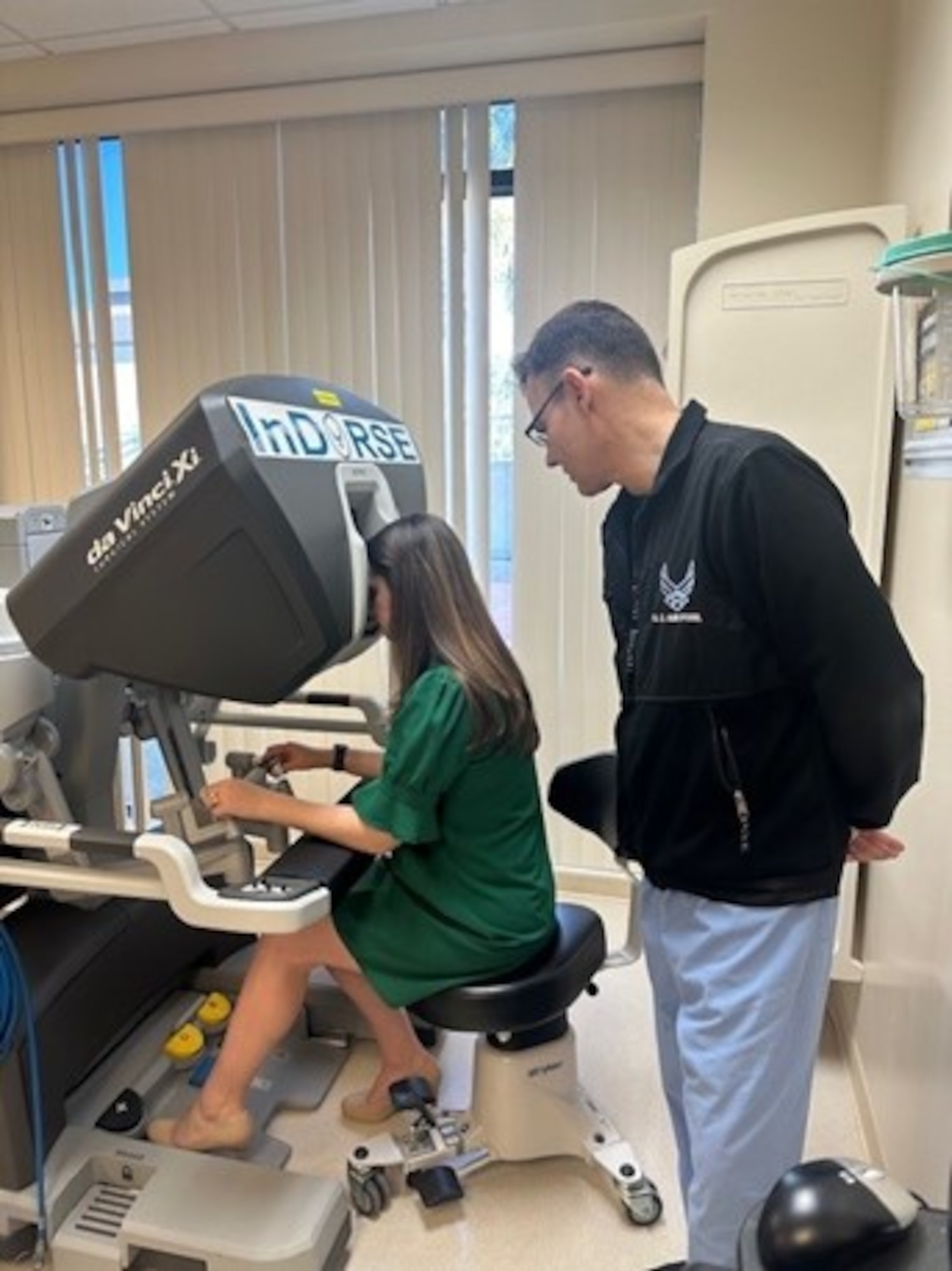 A serviceman in baby blue scrubs wearing a black hoodie with the Air Force logo on it stands over a woman in a green dress sitting down, operating machinery.