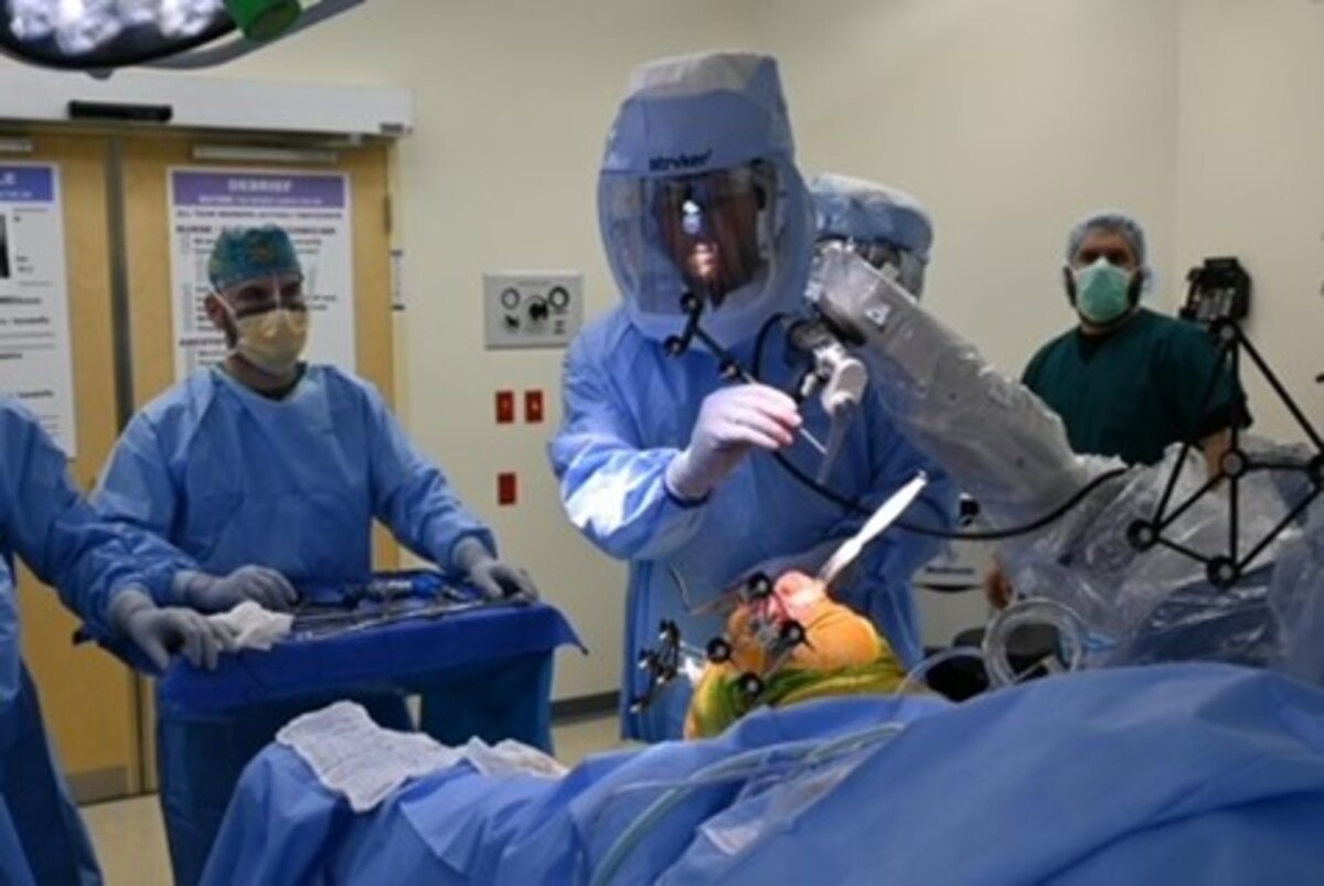 A man in surgical personal protective equipment operates on a body while surrounded by medical personnel in scrubs wearing regular masks.