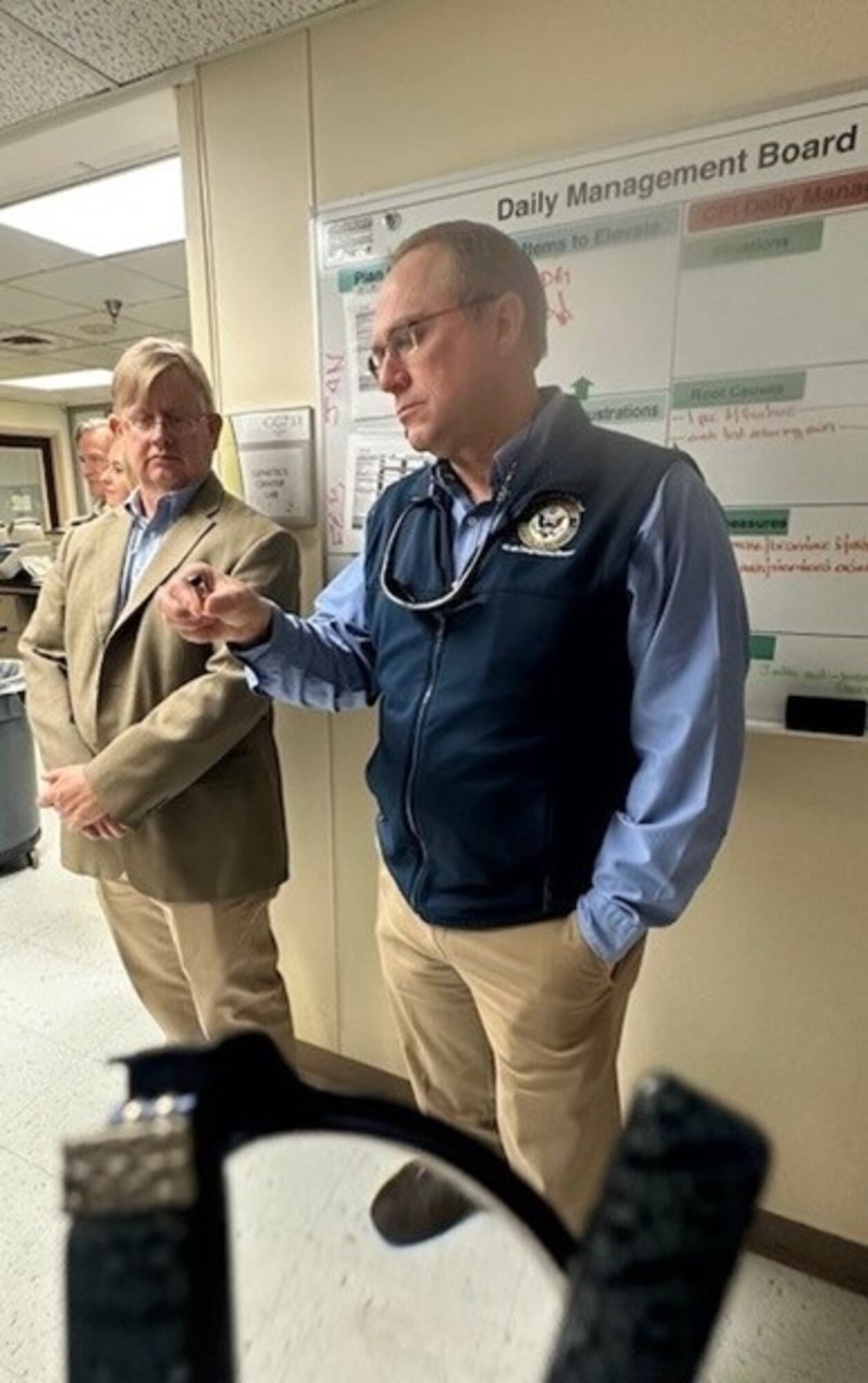 A man in khakis, wearing a baby blue long sleeve shirt with a navy blue vest looks at a glass vial alongside a man in a tan suit jacket also wearing kahkis. Both are standing in front of a white board.