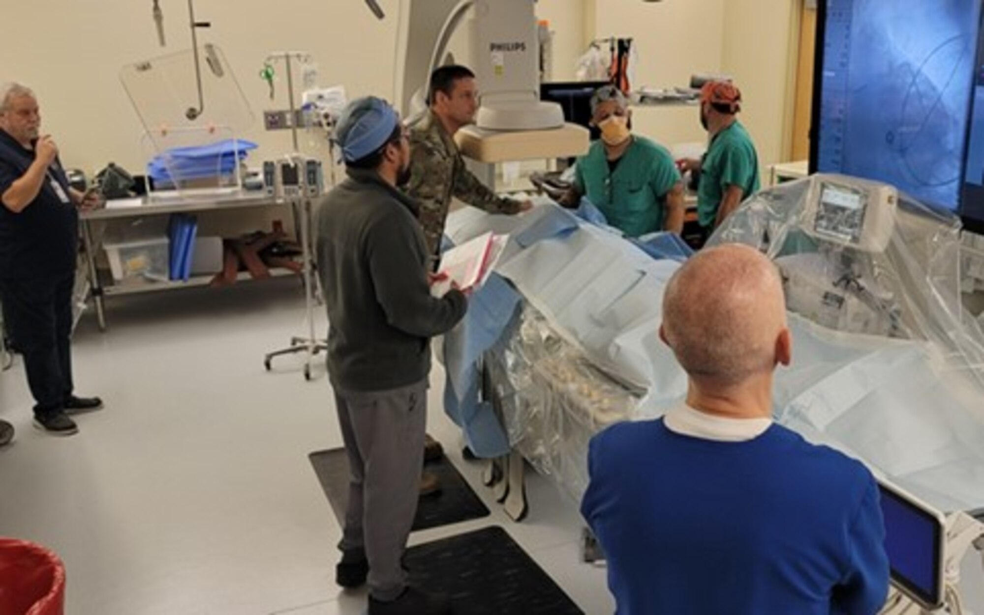 Medical personnel, wearing blue, green, or brown scrubs, surround a table while looking at a screen depicting an image of a vein.