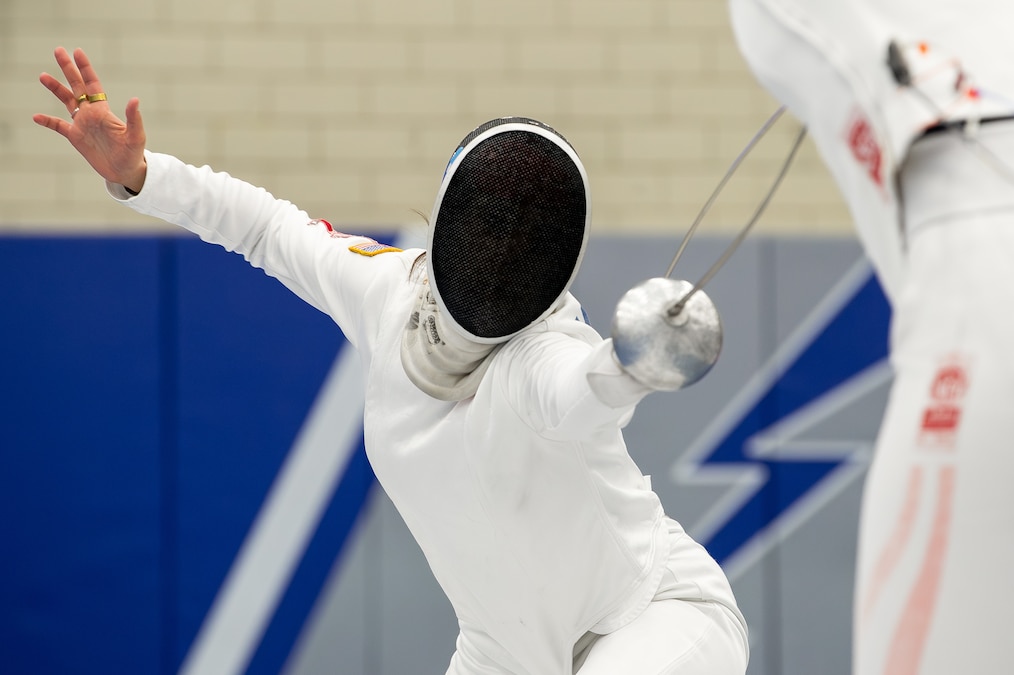 A fencer in full gear lifts one arm and aims the other with a sabre toward an opponent with a blue, white and gray wall in the background.