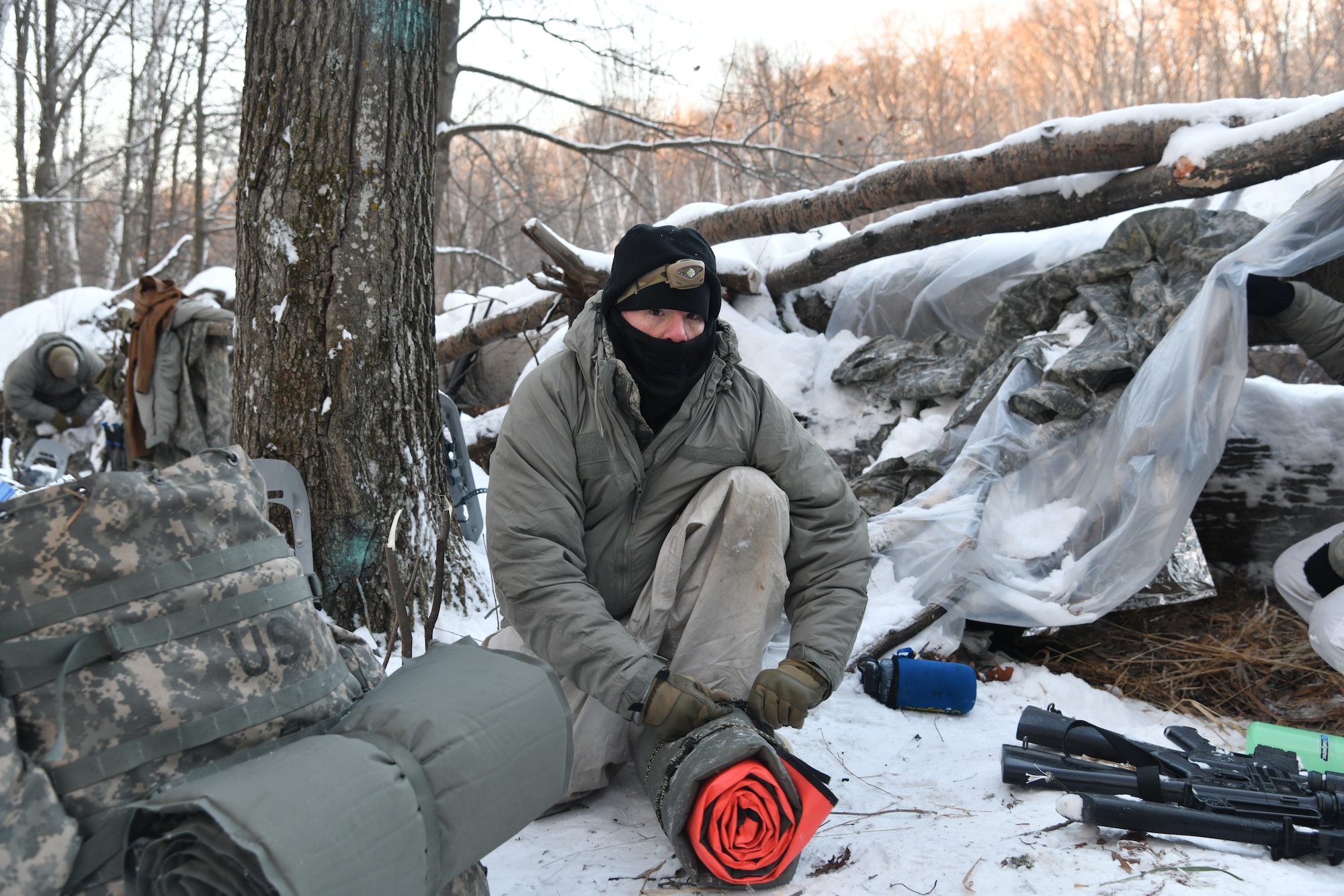 A man squats in the snowy woods while packing survival gear into a small travel size.