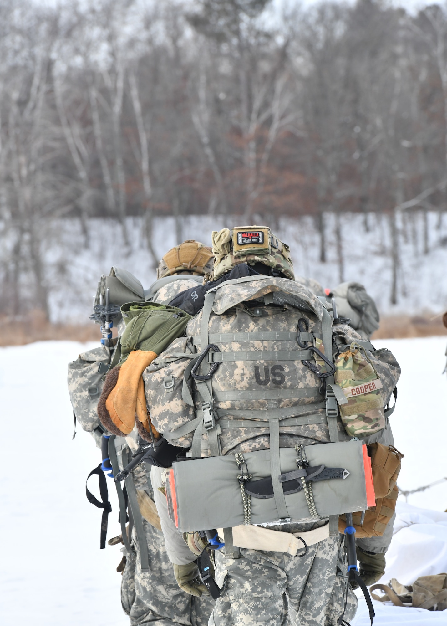 A man walks outdoors carrying a backpack with many survival supplies and attachments.