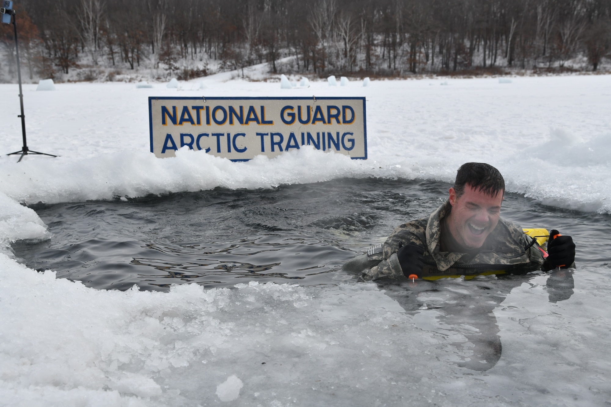 A man emerges from a icy pool carved into a snowy lake. A sign behind him read "National Guard Arctic Training."