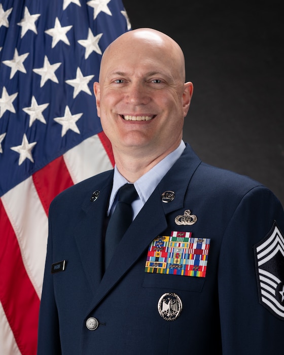Official Photo of CMSgt Jason Foster, USAF.  CMSgt Foster is wearing blue service dress in front of an American flag background.
