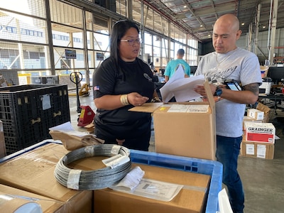 A man and a woman stand in a warehouse in front of boxes looking at papers.