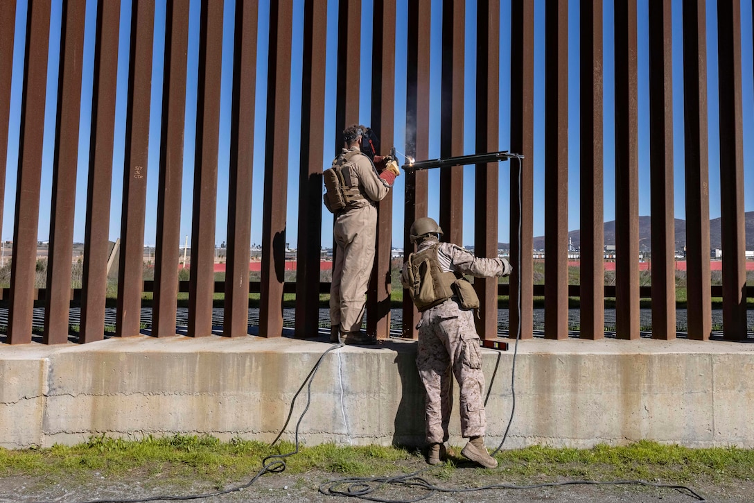 Two Marines weld a metal bar onto a wall of metal bars during the day.