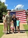 U.S. Army Soldier in uniform poses with wife, two daughters and son outside in front of an American Flag being held by two Soldiers in uniform