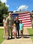 U.S. Army Soldier in uniform poses with wife, two daughters and son outside in front of an American Flag being held by two Soldiers in uniform