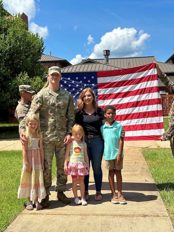 U.S. Army Soldier in uniform poses with wife, two daughters and son outside in front of an American Flag being held by two Soldiers in uniform