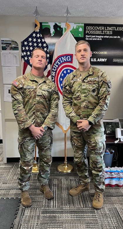 2 male U.S. Army Soldiers in uniform pose together in an office space in front of the American Flag and USAREC Flag