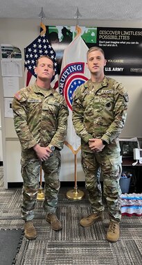 2 male U.S. Army Soldiers in uniform pose together in an office space in front of the American Flag and USAREC Flag