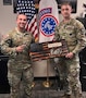 2 male U.S. Army Soldiers pose together with an American Flag plaque denoting NCO Of the Year