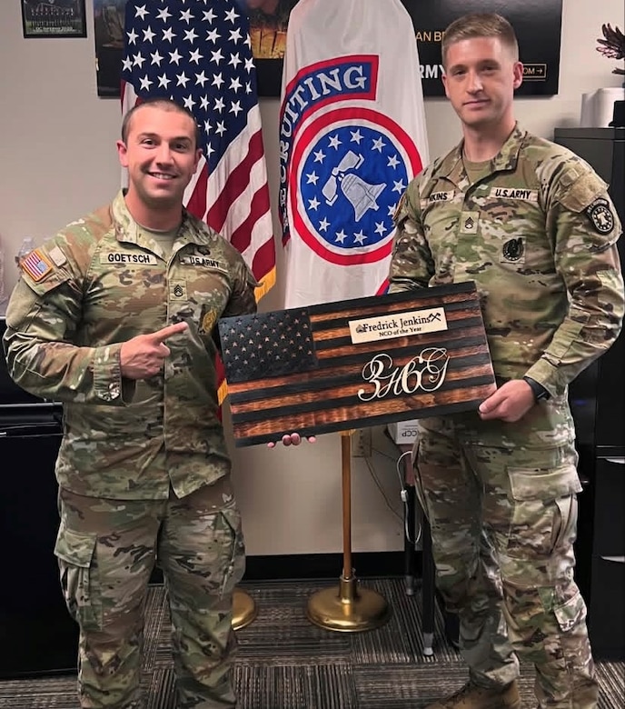 2 male U.S. Army Soldiers pose together with an American Flag plaque denoting NCO Of the Year