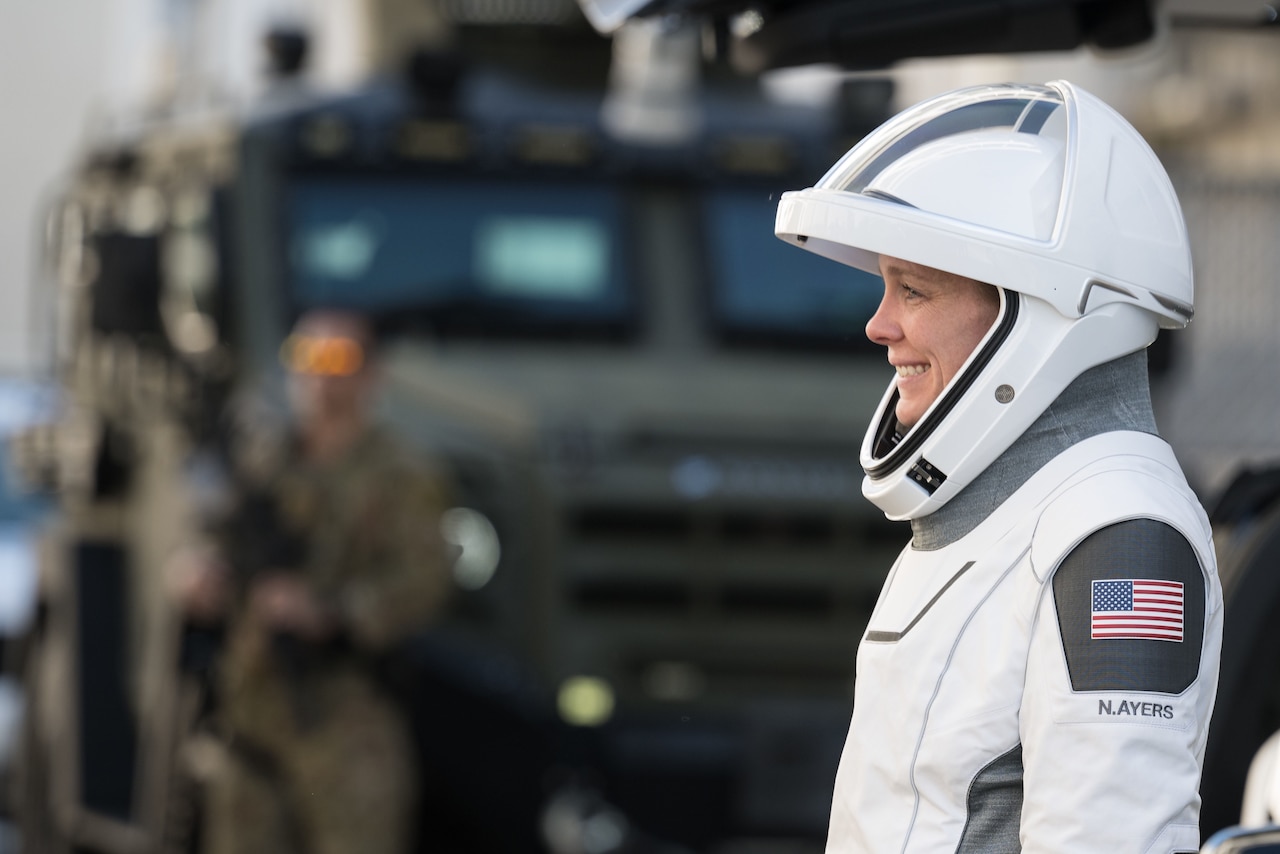 A woman in a white spacesuit smiles in front of a military vehicle.