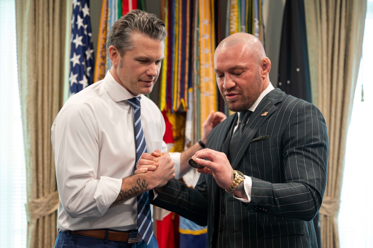Defense Secretary Pete Hegseth and another person clasp fists while examining a challenge coin in a room with flags in the background.