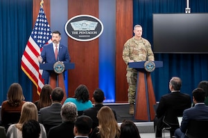 Two men, one in business attire and one in military camouflage, stand behind a pair of lecterns speaking to people seated and facing them.