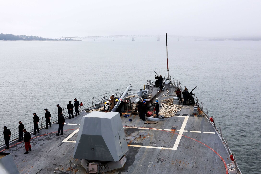 People wearing military uniforms line the edge of ship as it heads out to sea.