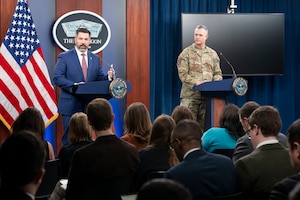 Two men, one in a military uniform, stand near each other behind lecterns. Behind them is an American flag and a sign that reads 