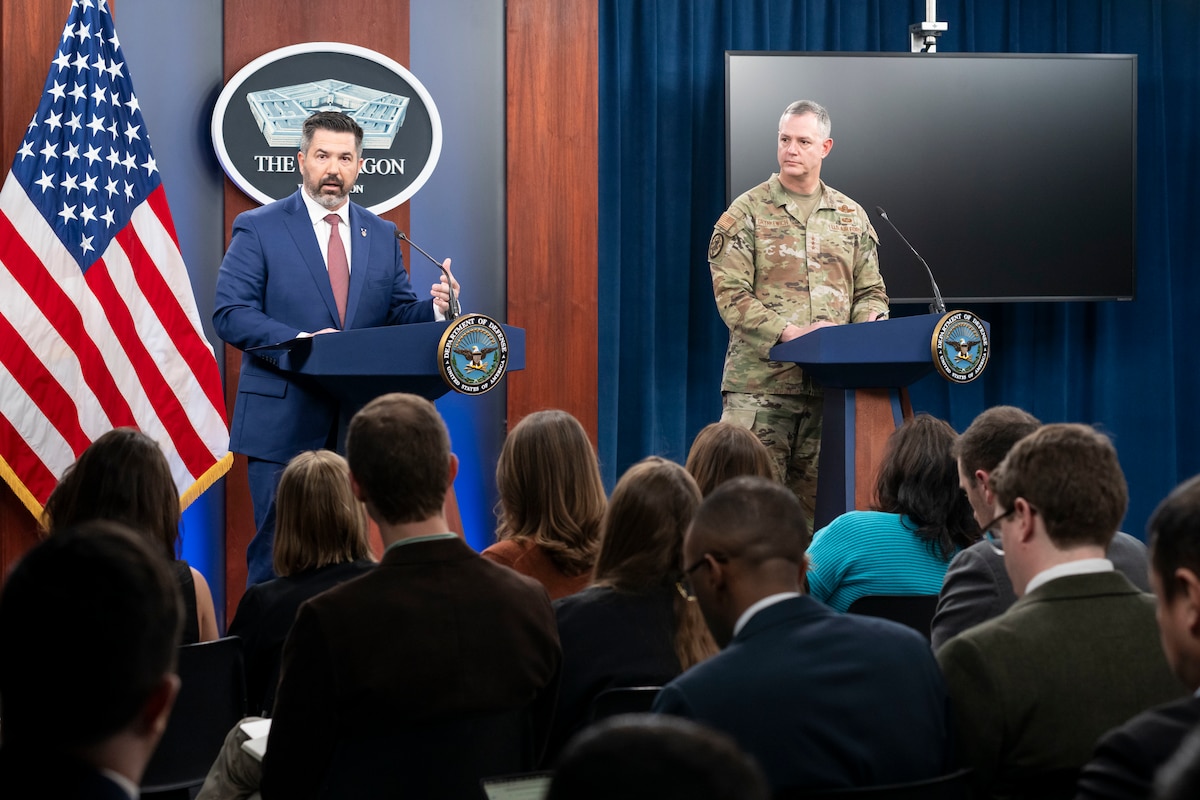 Two men, one in a military uniform, stand near each other behind lecterns. Behind them is an American flag and a sign that reads "The Pentagon – Washington."