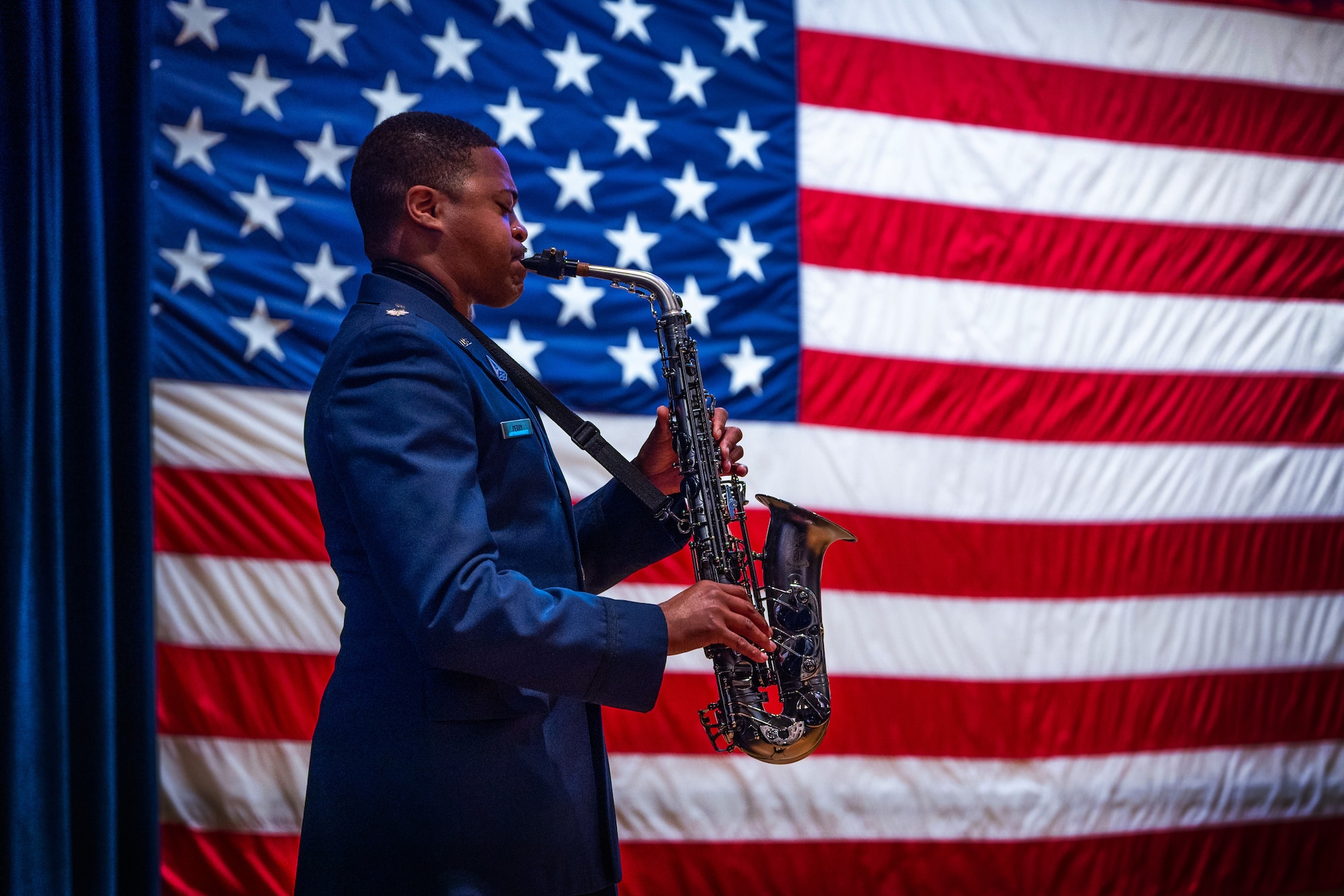 A man in uniform plays the saxophone in front of the United Stated flag.