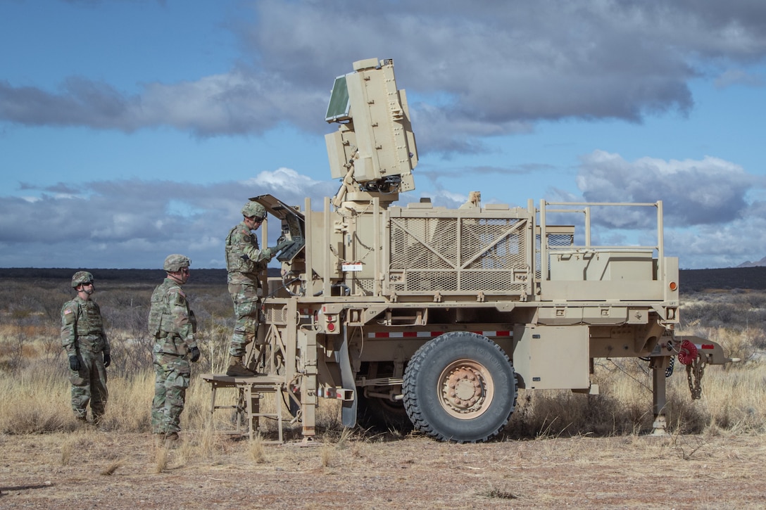 Three soldiers work on a large piece of military equipment outdoors on an arid field.