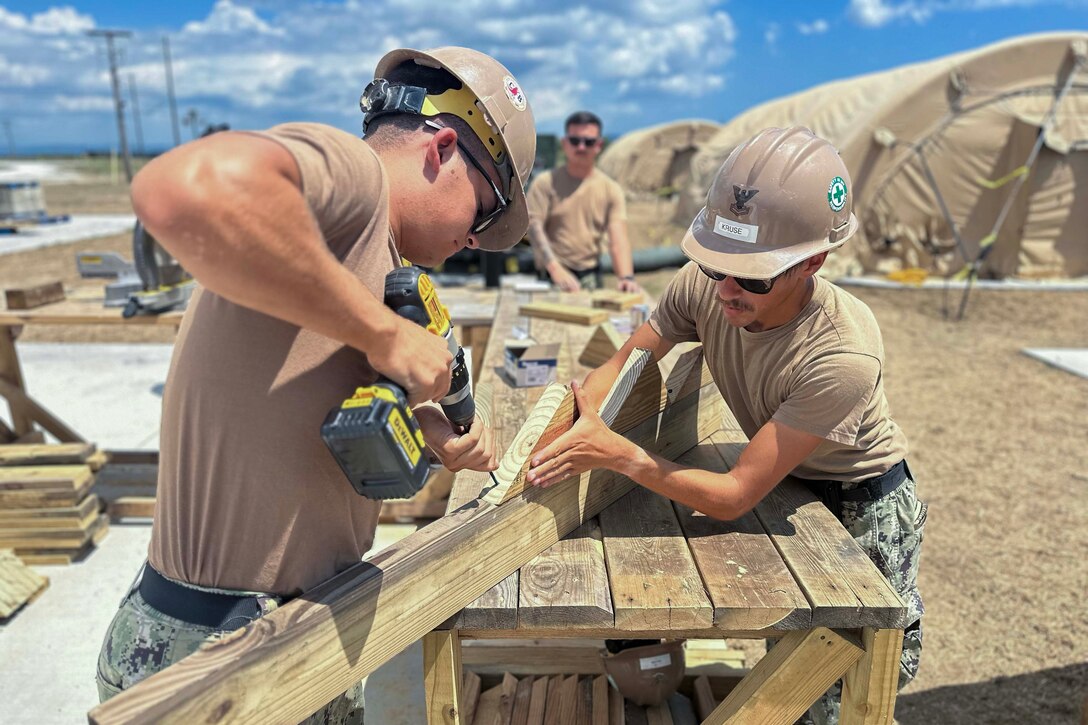 A sailor holds a wooden wedge to a plank while another sailor drills a hole in the wood.