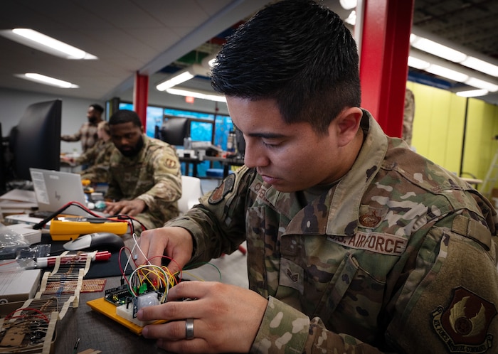 An Airman connects resistors to a circuit board with Airmen in the background working on circuit board projects.