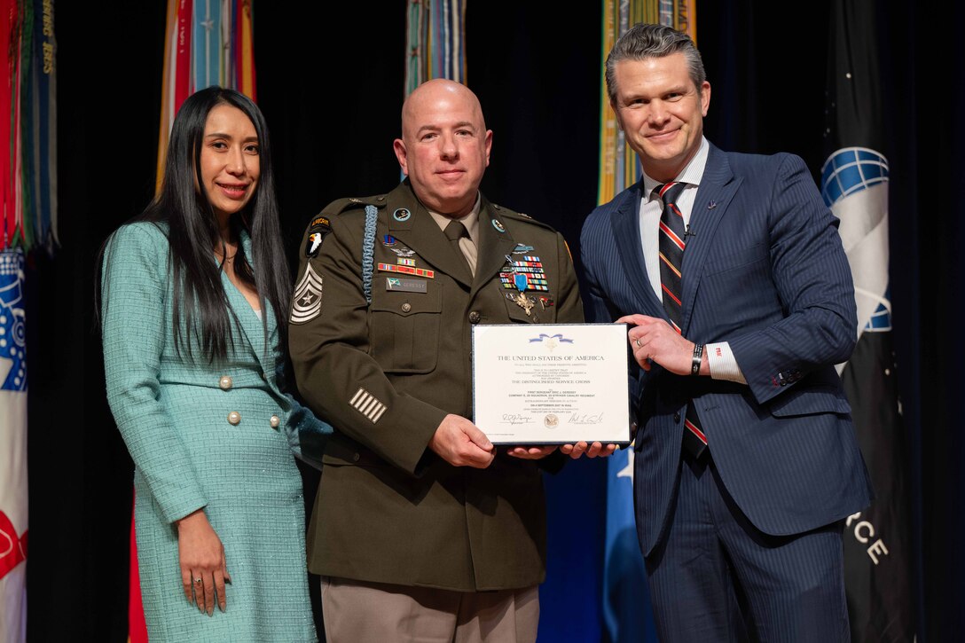 A former soldier holds an award together with a man wearing a suit. A woman stands on the other side.
