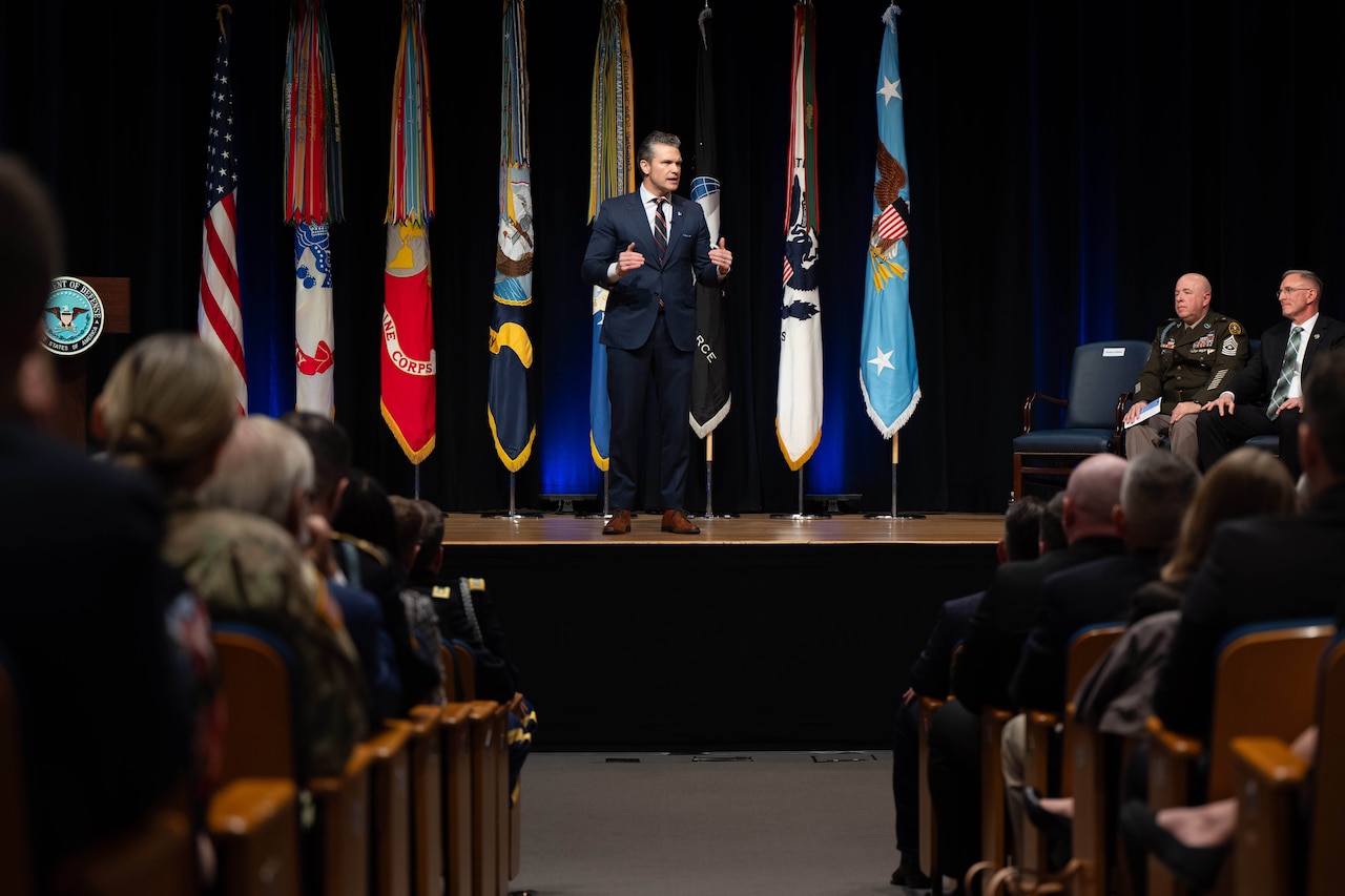 A man in a suit and tie speaks on stage to a crowded auditorium.