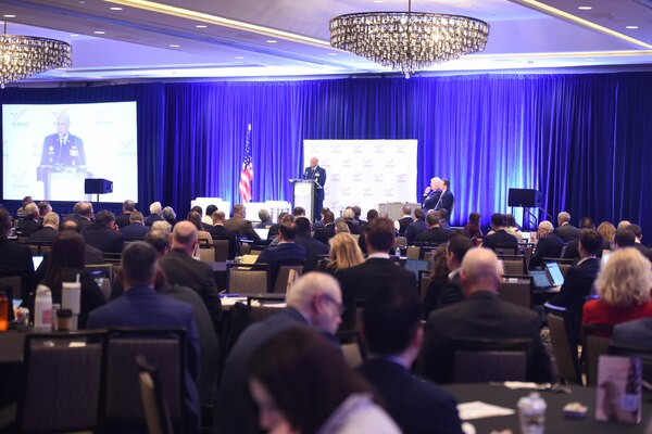 A man wearing a formal military uniform speaks at a podium to a room filled with people.