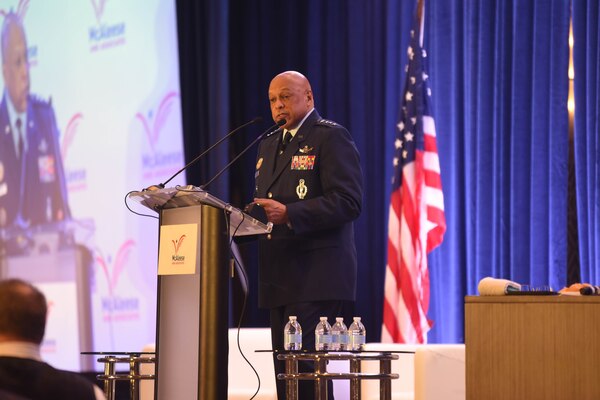 A man wearing a formal military uniform speaks at a podium to a room filled with people.