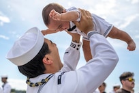 A sailor in white uniform holds up a smiling baby on a sunny day.