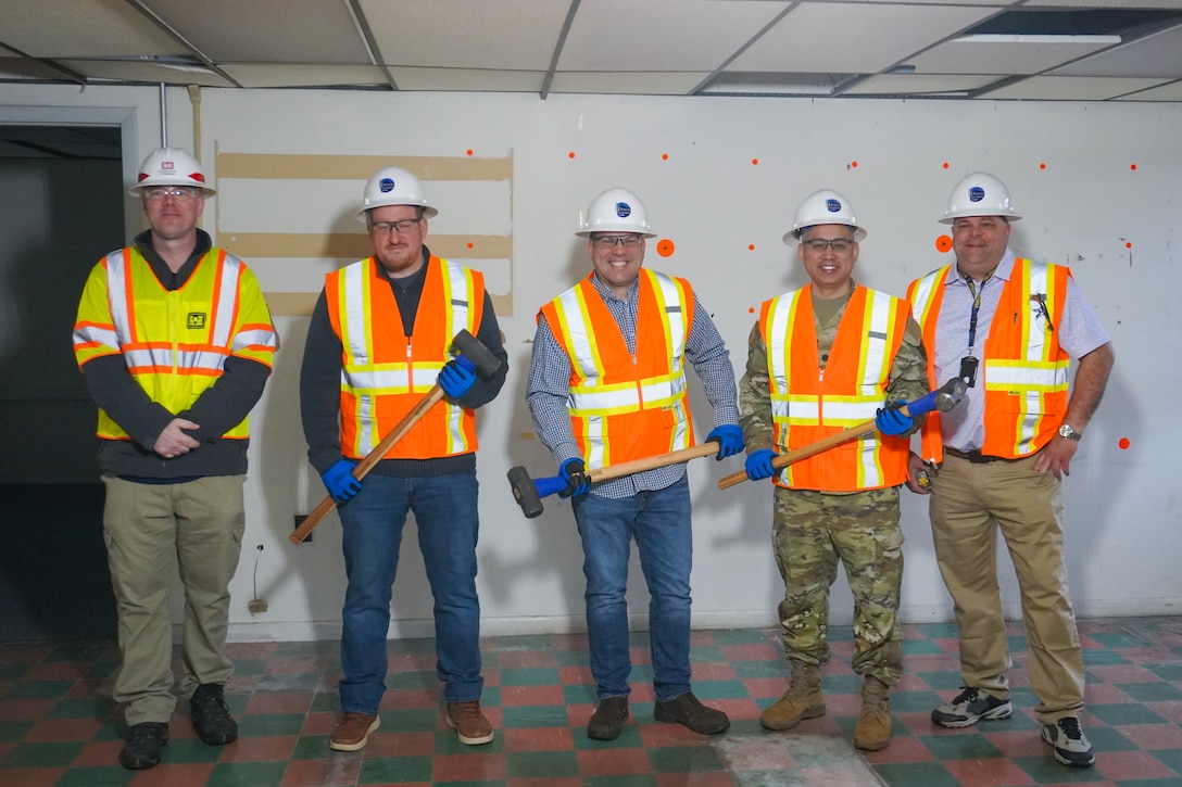 A group of five men wearing hard hats and reflective safety vests holding sledgehammers stand in front of a drywalled wall in a vacant room.
