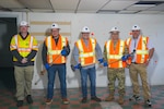 A group of five men wearing hard hats and reflective safety vests holding sledgehammers stand in front of a drywalled wall in a vacant room.