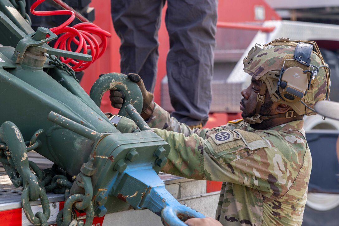 A paratrooper wearing combat gear attaches a winch on a equipment on the bed of a truck during the day.
