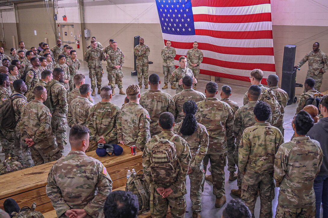 A group of soldiers gather around their commander in a gymnasium with a large American flag hanging from a wall.