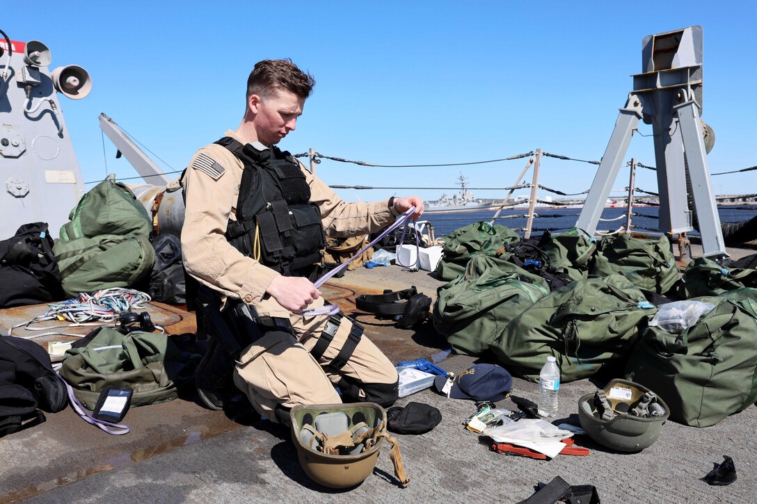 A sailor kneels down on the deck of a military ship and checks equipment strewn about.