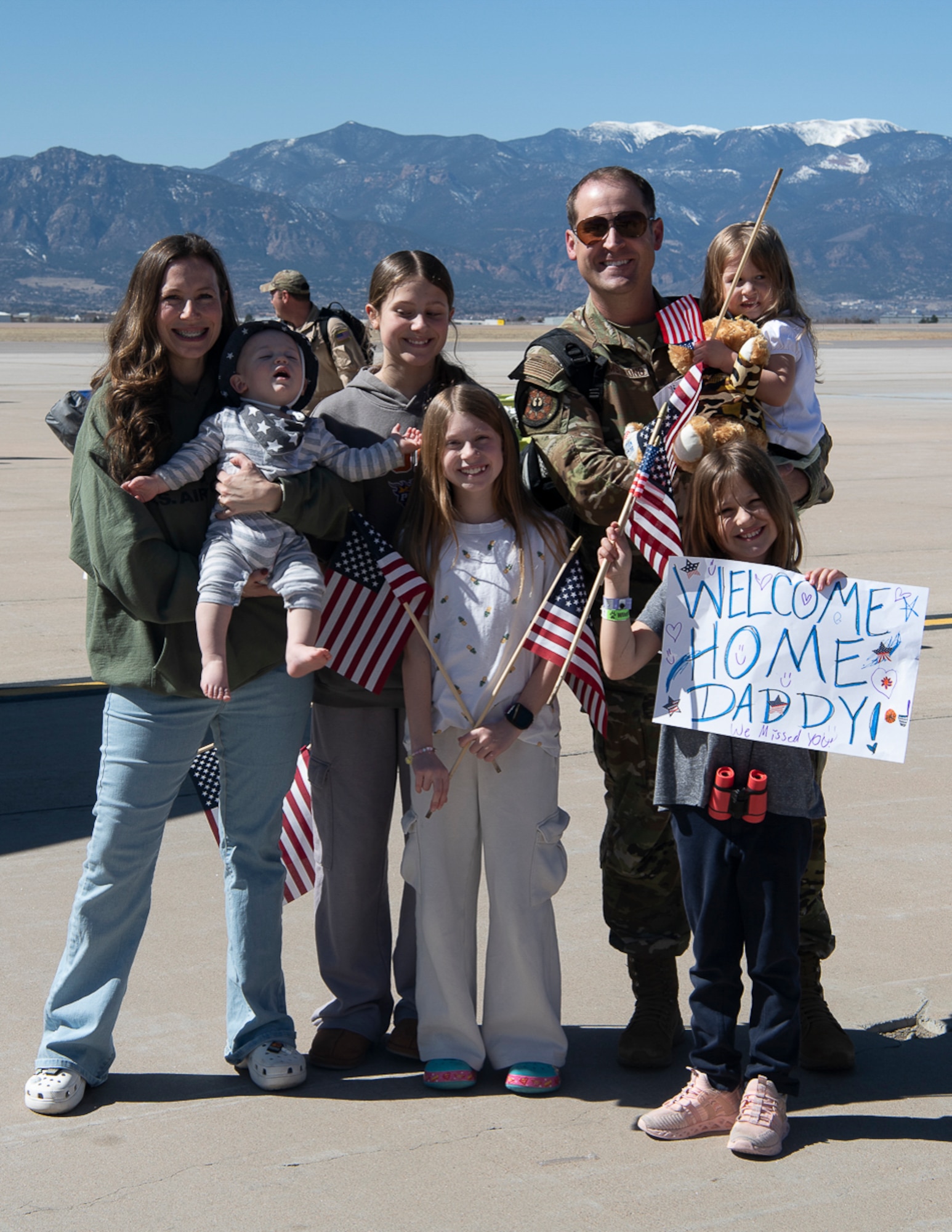 A family standing together with a child holding a sign that reads "WELCOME HOME DADDY!" on it.