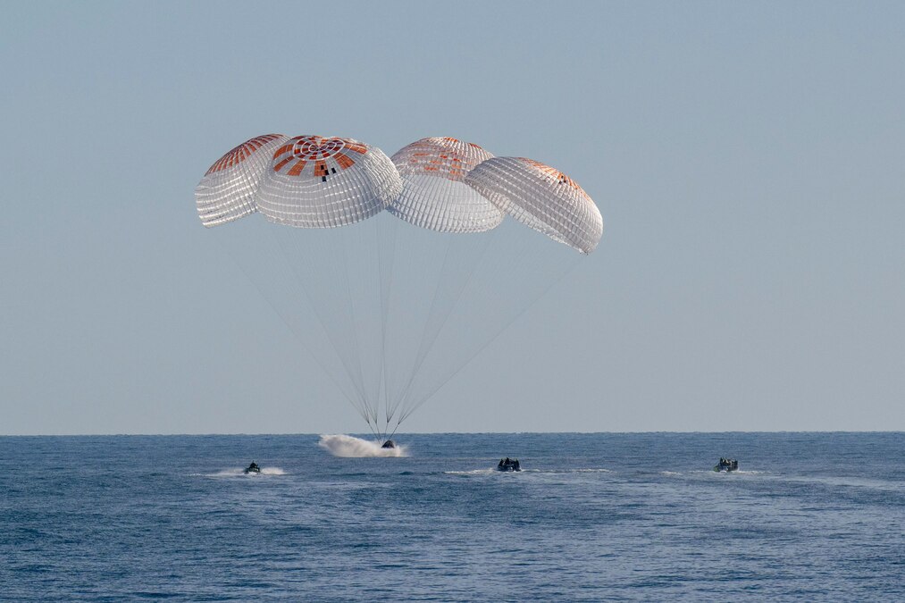 A spacecraft with four extended parachutes lands in the ocean while two small boats move towards the spacecraft.