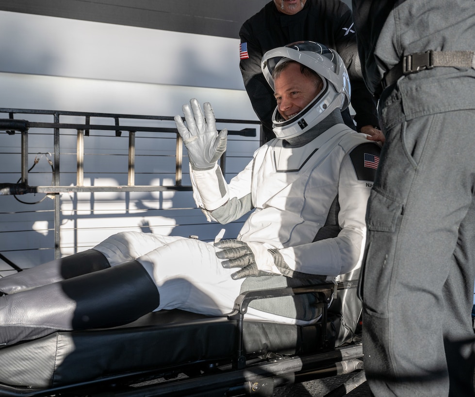 An astronaut in a white spacesuit waves while seated in a spacecraft.